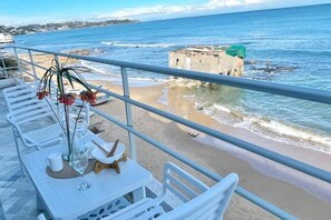 A balcony with a table set for dining, overlooking a beach with waves and a distant island. - L'appartement Pied Dans L'eau (La Marsa)