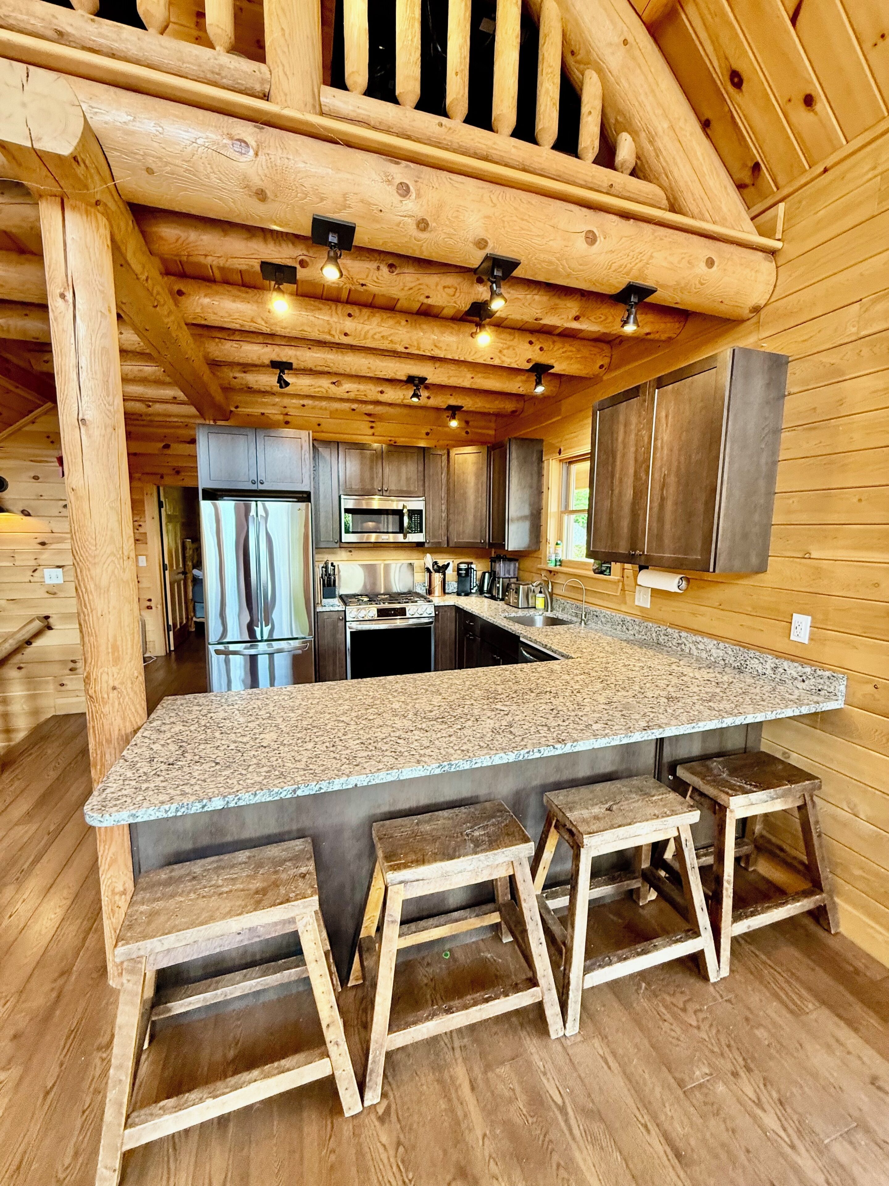 A kitchen with a granite countertop, wooden stools, stainless steel appliances, and a wooden ceiling.