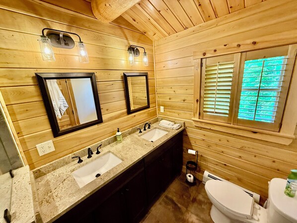 Une salle de bain rustique, avec des murs en bois, comprenant deux lavabos, des toilettes et une fenêtre équipée de stores.