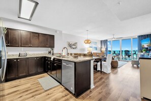 A modern kitchen with dark cabinets, a central island, and a dining area with a view. - 🌴Island Princess 301 - No Shoes Retreat | Gulf Views | Pool + Hot Tub🌴 (Fort Walton Beach)