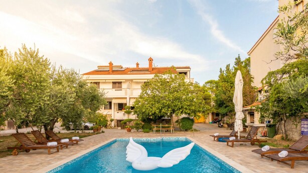Une piscine avec un cygne gonflable blanc, des chaises longues et, au fond, un bâtiment avec un toit recouvert de tuiles.