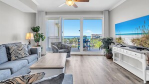Una sala de estar moderna con sofá, mesa de centro y televisor, un balcón con vistas a la playa y un ventilador de techo.