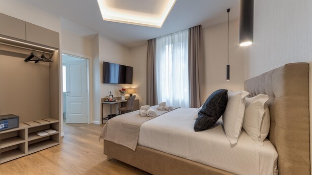 A modern hotel room with a large bed, a desk, a TV, and a walk-in closet. - GH Villa Poerio   Deluxe King Room (ROMA)