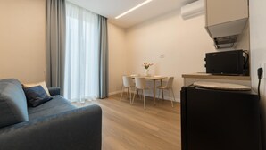 Ein modernes Wohnzimmer mit einer Couch, einem Esstisch mit Stühlen, einer kleinen Küchenzeile inklusive Kühlschrank und einem Fenster mit Vorhängen.
