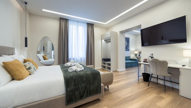 A modern hotel room with a bed, desk, chair, TV, and a large mirror. - GH Villa Poerio   Quadruple Room with Bath (ROMA)
