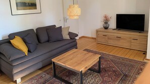 Un salon comprenant un canapé, une table basse, un téléviseur posé sur un meuble en bois, un luminaire suspendu et un tapis à motifs.