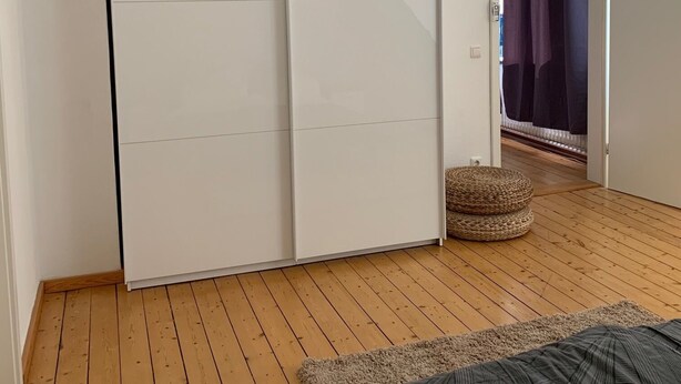 Une chambre avec un plancher en bois, un lit recouvert d’une couvre-lit à motifs, une armoire blanche et une porte donnant sur une autre pièce.