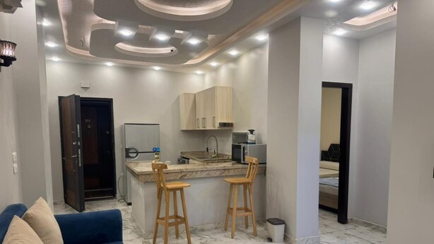 A modern living room with a blue sofa, a coffee table, and a kitchen area with wooden cabinets and a marble countertop. - Two bedroom apartment with Swimming Pool in the Intercontinental area, Hurghada (Red Sea Governorate)