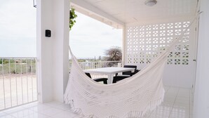 Una cadira d'hamaca blanca en una habitació amb taula i cadires blanques, una porta blanca amb un disseny de graella i vista a l'exterior.