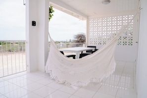 A white hammock chair in a room with a white table and chairs, a white door with a lattice design, and a view of the outdoors.