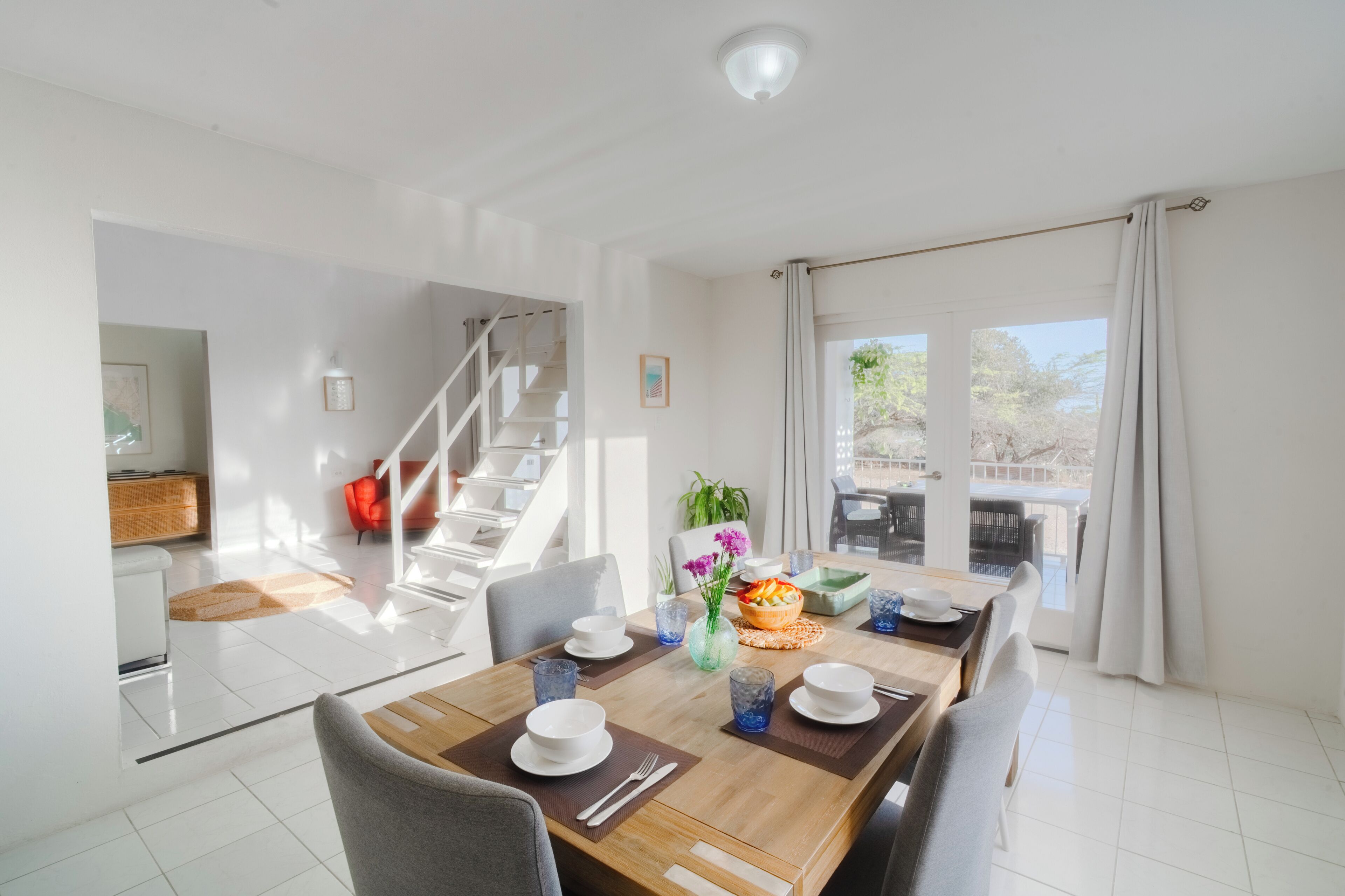 A bright dining area with a wooden table set for two, a staircase, and a view of the outdoors through large windows.