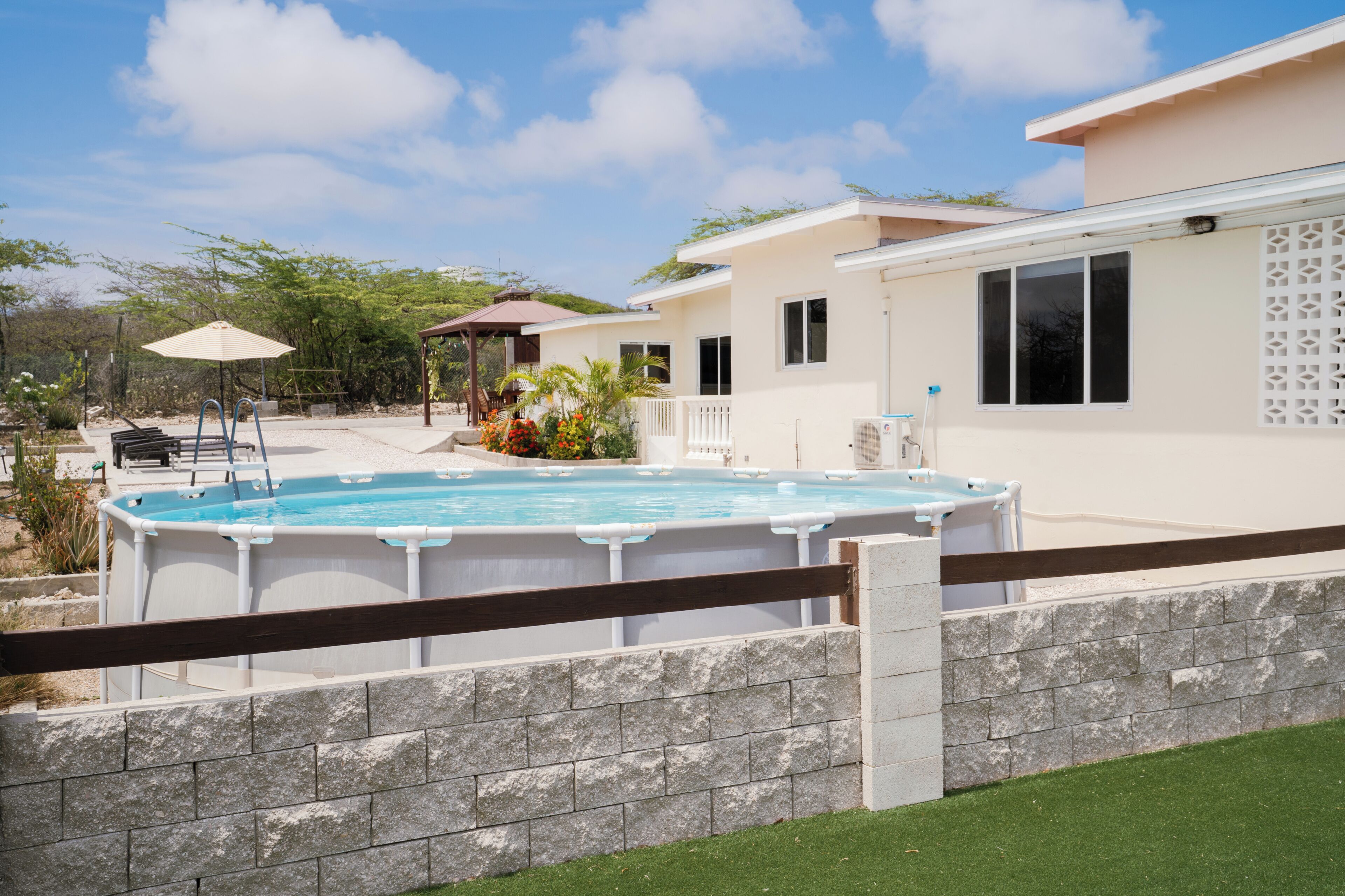 A swimming pool with a wooden deck, a covered seating area, and a house with a white exterior and a fenced yard.