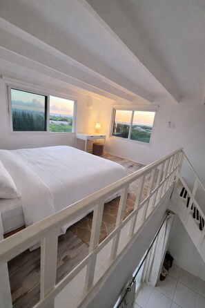 A bedroom with a large bed, a desk, and two windows offering a view of the landscape. - Villa on a Hill | Private Pool, Ocean Views, Aruba (Savaneta)