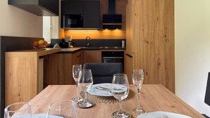 Une cuisine compacte dotée d’appareils électroménagers modernes, un coin repas avec une table en bois dressée pour deux personnes, et un espace salon confortable.