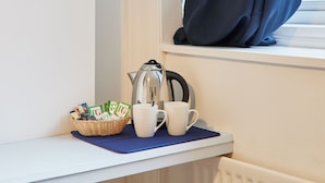 Een kitchenette met een waterkoker, twee mokken en een mandje theezakjes op een vensterbank.