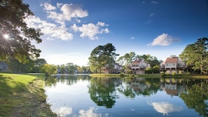 Une vue paisible sur un lac, avec le reflet des maisons et des arbres dans l’eau, un ciel dégagé et une surface d’eau calme.