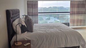 Une chambre à coucher avec un grand lit, une table de chevet, une lampe et une vue sur les immeubles par la fenêtre.