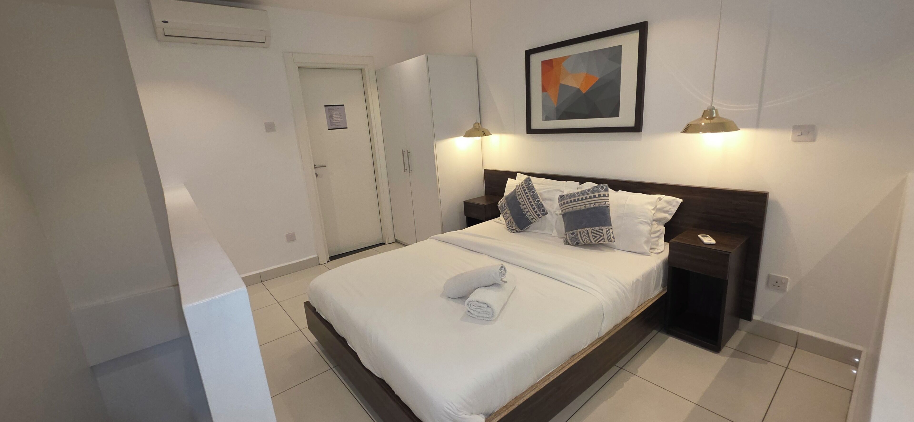 Una cama bien hecha con almohadas, una mesita de noche, un cuadro enmarcado en la pared y una puerta al fondo.