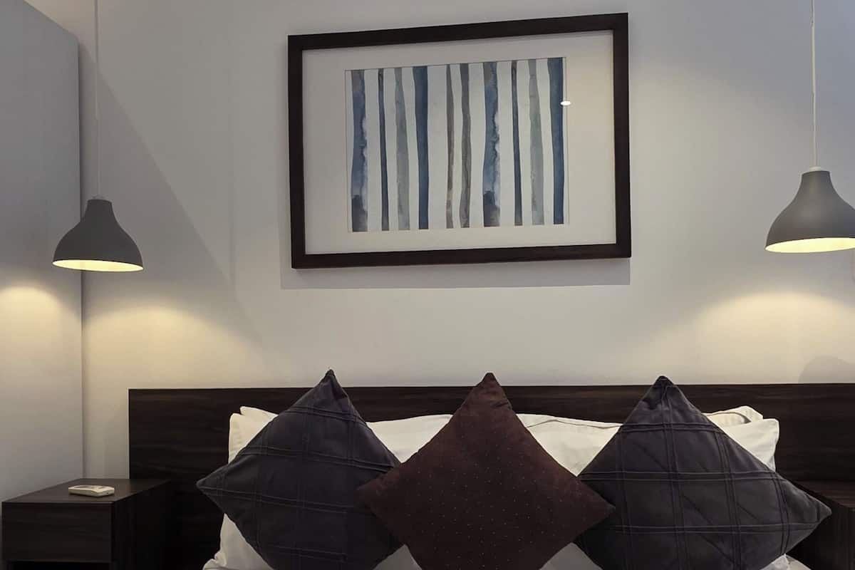 Una cama bien hecha con una obra enmarcada encima, dos lámparas de noche y un cabecero oscuro.
