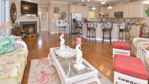 A living room with a fireplace, a television, and a kitchen area with bar stools. - Experience Oak Island living in "A Gift From Heaven," a 3BR water view retreat for 6. (Oak Island)