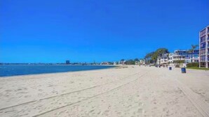 Ein Strand mit blauem Himmel, sandigem Ufer und Gebäuden im Hintergrund.