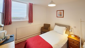 Una habitación de hotel con una cama, una silla, una mesita de noche con lámpara, un radiador, una ventana con cortinas y un cuadro en la pared.