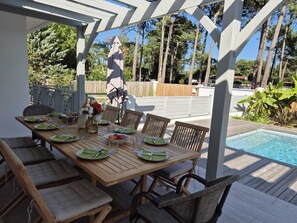 Ein Essbereich im Freien mit einem Holztisch, gedeckt für vier Personen, umgeben von Korbstühlen, unter einer weißen Pergola. Im Hintergrund ist ein Schwimmbecken zu sehen.