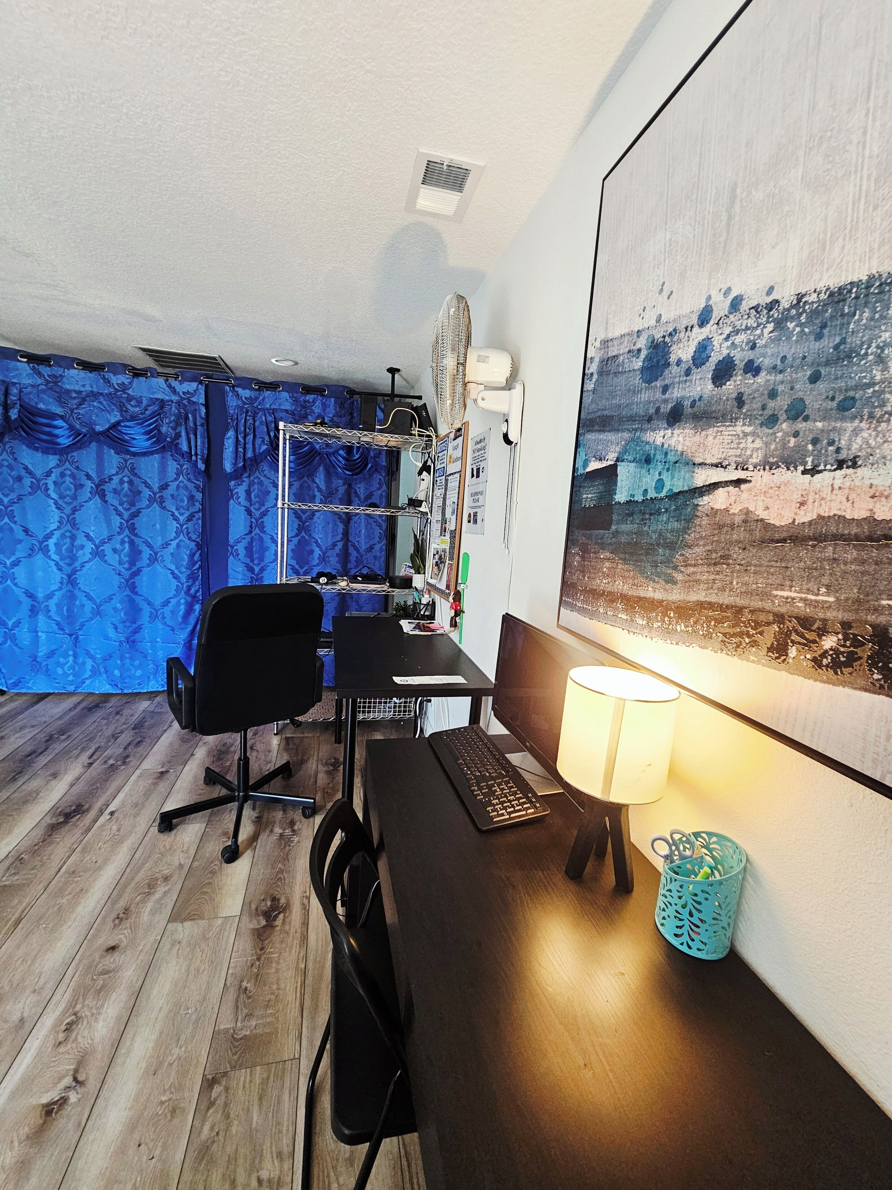 Een kamer met een bureau, stoel, computer, lamp en een ventilator aan de muur.
