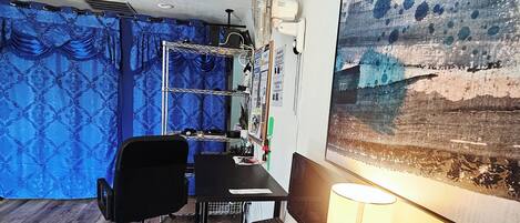 Een kamer met een bureau, stoel, computer, lamp en een ventilator aan de muur.