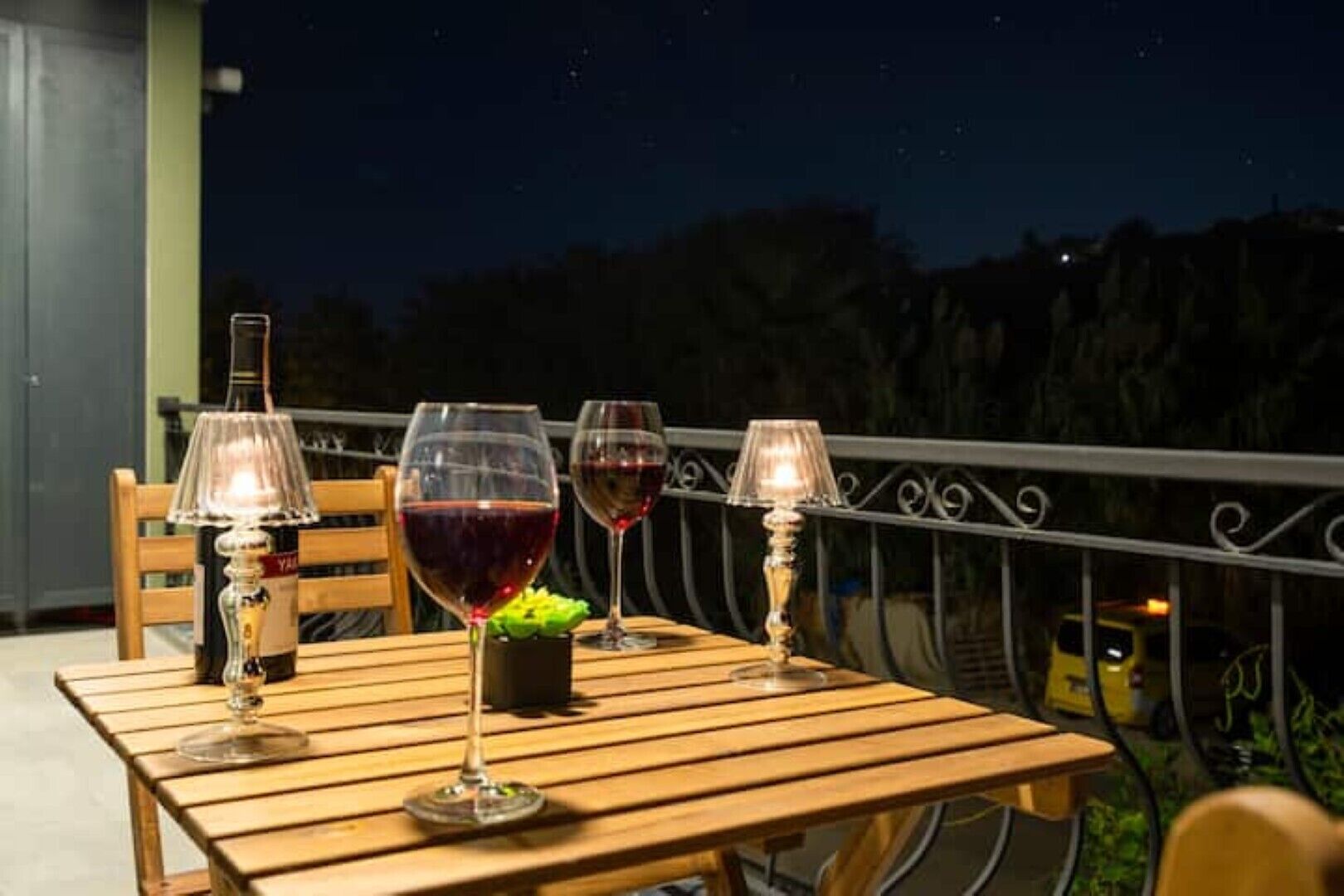Ein Holztisch mit drei Weingläsern und einer Flasche, einer Lampe und einer kleinen Pflanze auf einem Balkon bei Nacht.