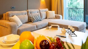 Une chambre d’hôtel avec un canapé, une table à manger avec des fruits, un tableau et une vue sur la verdure.