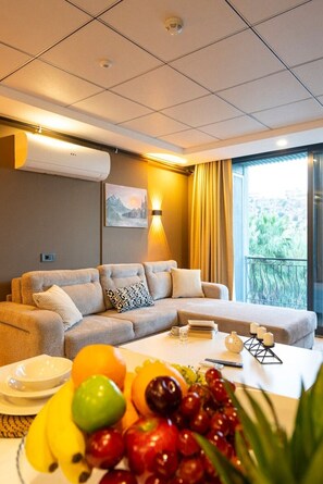 Ein Hotelzimmer mit einer Couch, einem Esstisch mit Obst, einem Gemälde und Blick auf Grünflächen.
