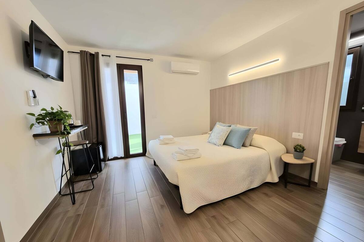 A modern bedroom with a bed, a TV, a desk with a plant, and a sliding door to a balcony.