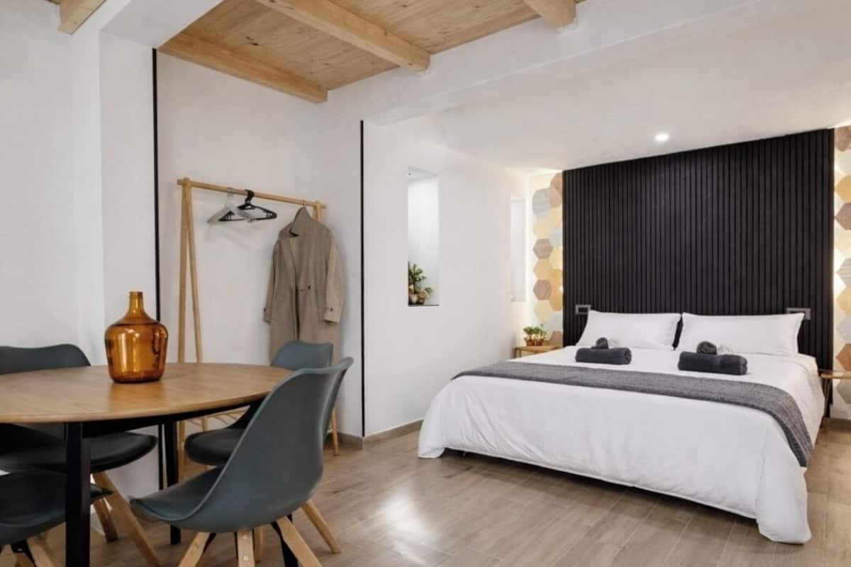 A modern room with a wooden ceiling, a bed with white bedding, a round table with chairs, and a coat rack with clothes.