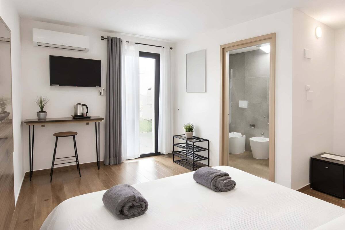 A modern hotel room with a large bed, a desk, a television, a mirror, and a bathroom visible through an open door.