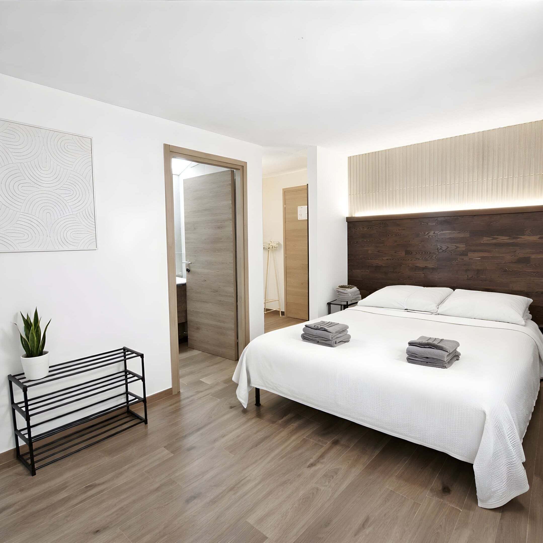A modern bedroom with a wooden floor, a bed with white bedding, a bedside table, a wall-mounted light, and a plant.