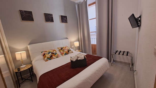 A hotel room with a bed, two bedside lamps, a TV mounted on the wall, and a window with curtains. - Nest Style Navas (Granada)