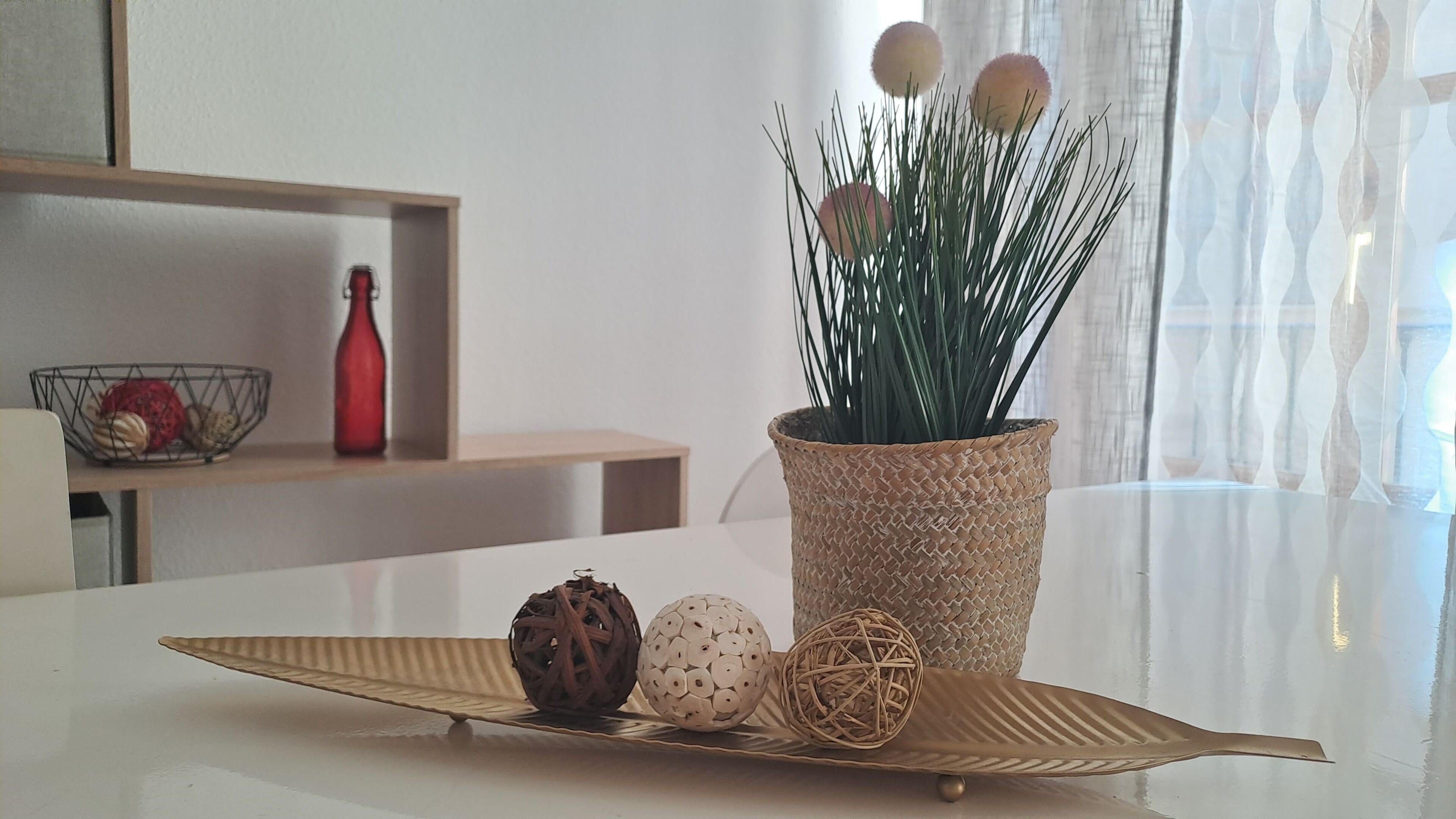 A table with a woven pot containing green plants, a decorative plate with three objects, and a red bottle on a shelf.