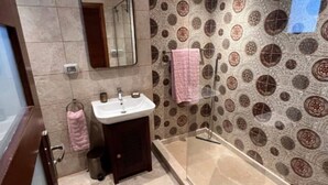 Baño con ducha de vidrio, lavamanos, espejo, azulejos estampados y una alfombra azul.