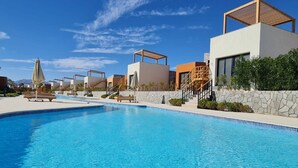 Una piscina junto a edificios modernos, un muro de piedra y un cielo azul despejado.