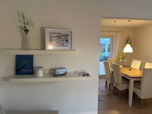 Un comedor con mesa y sillas de madera, un cuadro enmarcado en la pared, un estante pequeño con un libro y un jarrón de flores.