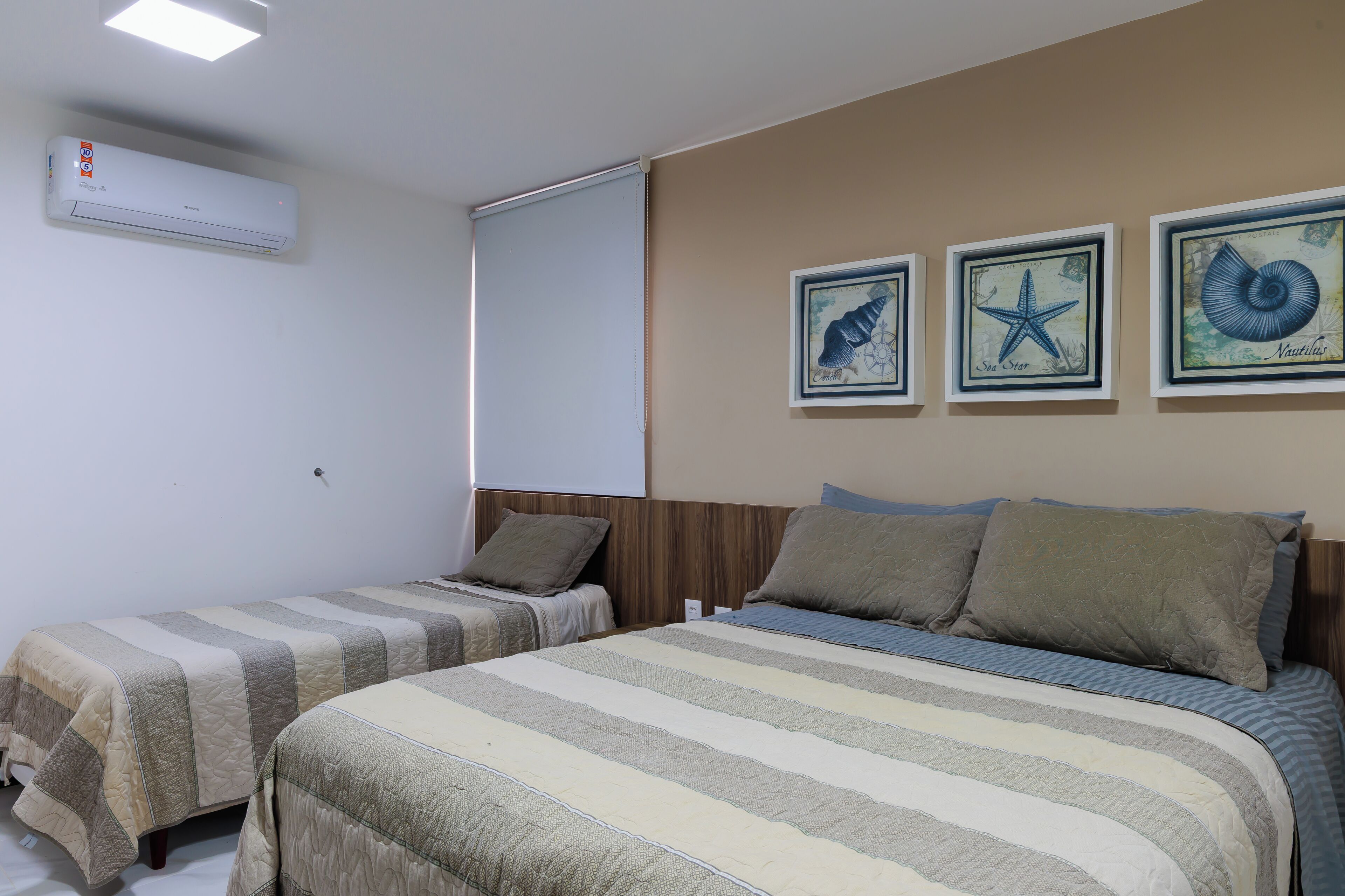 A hotel room with two beds, a ceiling fan, a wall-mounted air conditioner, and three framed nautical-themed pictures.