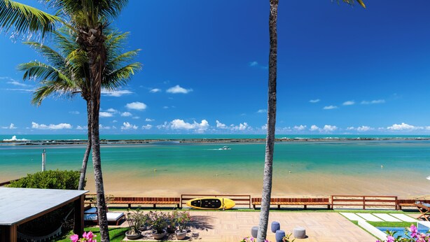 A beach view with a wooden deck, a yellow kayak, and palm trees. - Casa Manaua: beachfront property with incredible views in Barra de São Miguel (Barra de São Miguel)