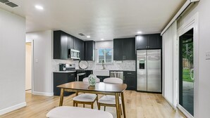 Uma cozinha moderna com armários escuros, eletrodomésticos de aço inoxidável, painel de fundo de mármore e um espaço de jantar com mesa e cadeiras de madeira.