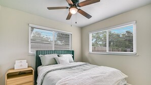 Um quarto com uma cama, uma mesa de cabeceira, um ventilador de teto e duas janelas com persianas.