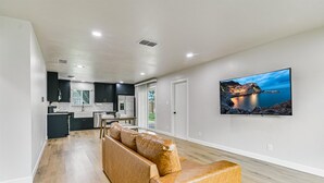Uma sala de estar moderna com um sofá de couro, uma TV de tela plana na parede e uma cozinha ao fundo.