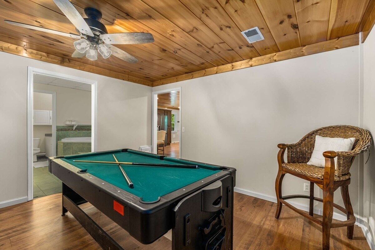 Une table de billard avec des queues, un ventilateur de plafond et un fauteuil en osier dans une pièce au plancher en bois et aux murs blancs.