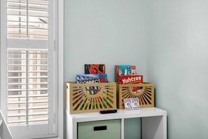 A room with a window with blinds, a white desk with two wooden crates containing board games, and a green cabinet below.