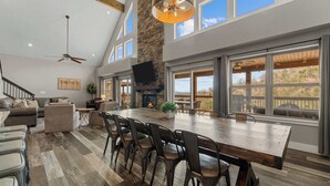 Amplia sala de estar con un gran comedor de madera, múltiples sillas, chimenea y ventanas grandes que ofrecen vistas al exterior.