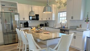 A modern kitchen with white cabinetry, a stainless steel refrigerator, a microwave, and a large island with bar stools. - Oceanview Key Rest on Oak Island: Spacious 6BR, 5BA home for 15 guests with pool. (Oak Island)